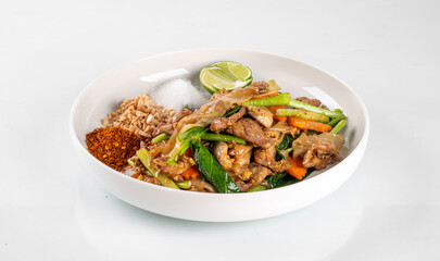 A delicious bowl of Thai stir-fried chicken with vegetables, served with rice, lime, and chili sauce on the side.