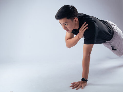 An Asian man holding a plank position while touching his shoulder with one hand, demonstrating balance and core strength. He is wearing a black t-shirt against a white background