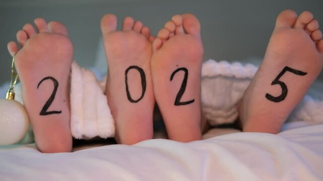 Close-up of two children's bare feet sticking out from under white blanket on bed with 2025 written on it. Winter New Year concept. Place for text.