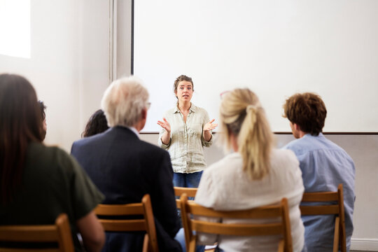 Confident businesswoman gesturing while giving presentation to coworkers against blank projection screen at office
