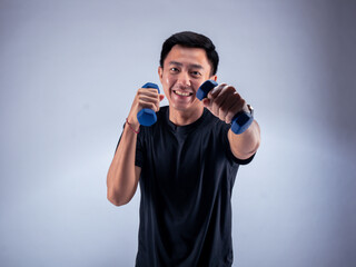 An Asian man smiling while performing a punch motion with blue dumbbells in both hands. He is wearing a black t-shirt and stands against a white background