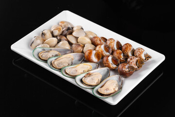 A variety of fresh and raw shellfish including clams, snails, and mussels served on a rectangular white plate against a black background.