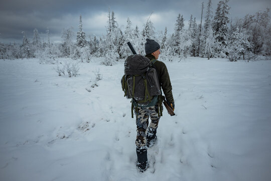 Rear view of hunter carrying rifle and backpack while walking on snow covered field