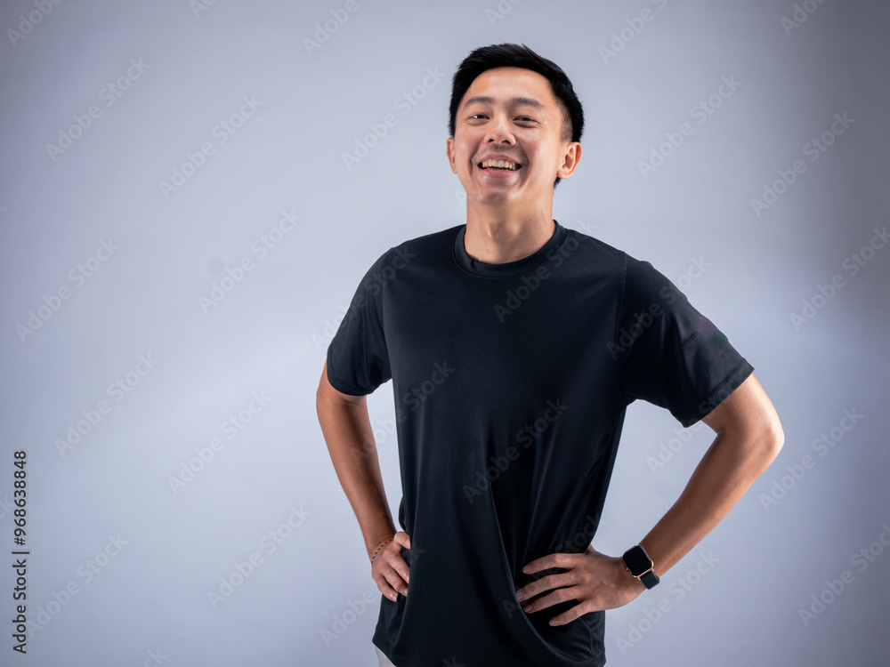 Wall mural An Asian man stands with hands on hips, smiling confidently, wearing a black t-shirt and gray shorts. He poses against a white background in a fitness studio, showcasing a relaxed demeanor.