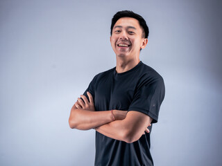 Fototapeta premium An Asian man standing confidently with arms crossed, smiling while wearing a black t-shirt. He stands against a white background in a fitness studio, showcasing a positive and relaxed attitude