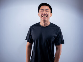 An Asian man smiling warmly while standing in a relaxed pose, wearing a black t-shirt. He is...