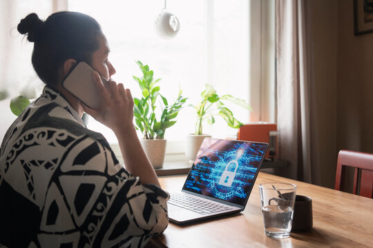 Woman talking on smart phone with encrypted laptop on table at home