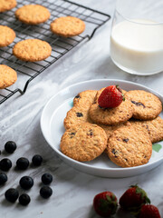 A plate of freshly baked oatmeal raisin cookies garnished with a strawberry, accompanied by a glass of milk and scattered blueberries, set against a light, natural background for a cozy snack scene.