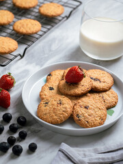 A plate of freshly baked oatmeal raisin cookies garnished with a strawberry, accompanied by a glass of milk and scattered blueberries, set against a light, natural background for a cozy snack scene.