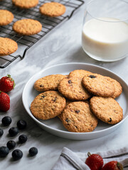 A plate of freshly baked oatmeal raisin cookies garnished with a strawberry, accompanied by a glass of milk and scattered blueberries, set against a light, natural background for a cozy snack scene.