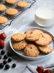 A plate of freshly baked oatmeal raisin cookies garnished with a strawberry, accompanied by a glass of milk and scattered blueberries, set against a light, natural background for a cozy snack scene.
