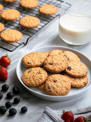 A plate of freshly baked oatmeal raisin cookies garnished with a strawberry, accompanied by a glass of milk and scattered blueberries, set against a light, natural background for a cozy snack scene.