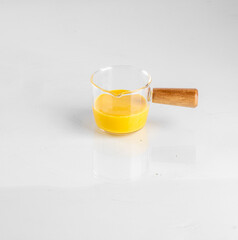 Clear glass measuring cup with a wooden handle, containing orange liquid on a white background, modern minimalistic cooking concept.