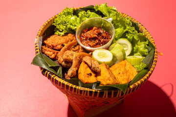 A traditional Indonesian meal served in a woven bamboo basket with fried fish, tofu, cucumber slices, lettuce, and spicy chili paste on banana leaves, creating a vibrant and appetizing presentation
