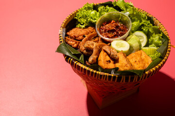 A traditional Indonesian meal served in a woven bamboo basket with fried fish, tofu, cucumber slices, lettuce, and spicy chili paste on banana leaves, creating a vibrant and appetizing presentation