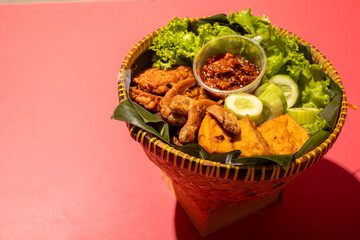 A traditional Indonesian meal served in a woven bamboo basket with fried fish, tofu, cucumber slices, lettuce, and spicy chili paste on banana leaves, creating a vibrant and appetizing presentation