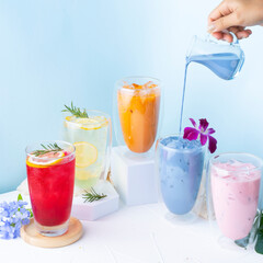 Colorful refreshing drinks with flowers and a hand pouring blue milk. Perfect summer beverage setup on a light blue background.