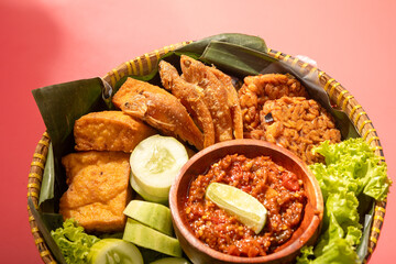 A traditional Indonesian meal served in a woven bamboo basket with fried fish, tofu, cucumber slices, lettuce, and spicy chili paste on banana leaves, creating a vibrant and appetizing presentation