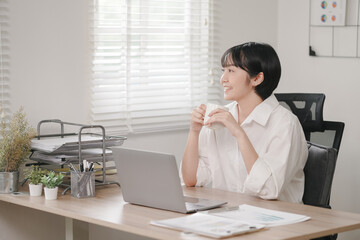 A woman is sitting at a desk with a laptop and a cup of coffee. She is smiling and she is enjoying her work