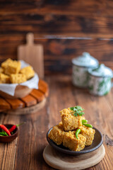 A serving of crispy fried tofu cubes garnished with green chili, presented in a rustic bowl on a wooden table. The background features a blurred bowl of tofu and traditional ceramic containers