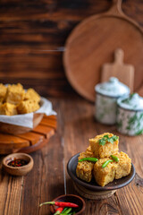 A serving of crispy fried tofu cubes garnished with green chili, presented in a rustic bowl on a wooden table. The background features a blurred bowl of tofu and traditional ceramic containers