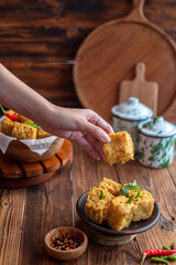 A hand picking up a crispy fried tofu cube from a rustic bowl, served with green chili and soy-based dipping sauce. The scene is set on a wooden table with containers in the background