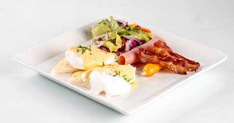 Delicious breakfast plate with poached eggs, crispy bacon, fresh salad, and avocado on a white square plate.