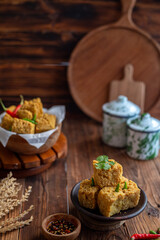A serving of crispy fried tofu cubes garnished with green chili, presented in a rustic bowl on a wooden table. The background features a blurred bowl of tofu and traditional ceramic containers
