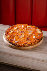 Delicious cheese and ham pizza on a wooden board, placed on a marble table against a red cushioned background.