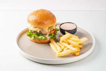 Delicious cheeseburger with lettuce, tomato, and fries served with dipping sauce on a gray plate, on a white background.