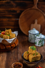 A serving of crispy fried tofu cubes garnished with green chili, presented in a rustic bowl on a wooden table. The background features a blurred bowl of tofu and traditional ceramic containers