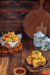 A serving of crispy fried tofu cubes garnished with green chili, presented in a rustic bowl on a wooden table. The background features a blurred bowl of tofu and traditional ceramic containers