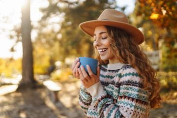 A young woman enjoys her coffee by the serene lakeshore during a sunny autumn afternoon surrounded by vibrant trees. Аutumn landscape near the river. The concept of travel, relax.