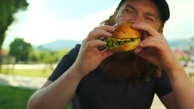 Bearded glutton man eats meat burger with pleasure, bites cheeseburger with his mouth in fast food restaurant outdoors.