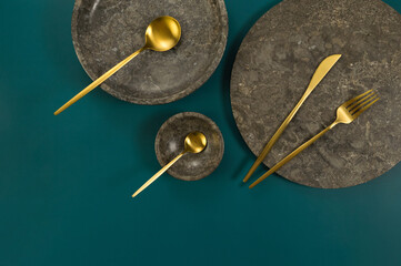 Top view of brown marble tray, plate, bowl and gold cutlery on dark green background. Food presentation, dishes, stone tray. Copy space, flat lay.