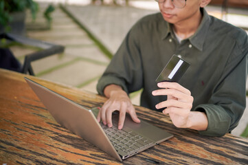 Young Asian man working on his laptop while talking on mobile phone and holding a credit card. Man trying to online purchasing with card