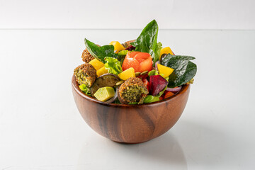 Fresh, colorful salad bowl with falafel, avocado, mango, spinach, and vegetables in a wooden bowl on a white background.