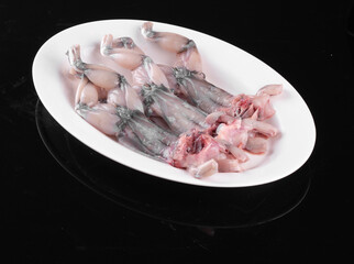 Fresh raw frog legs on a white plate, ready for cooking, isolated on a black background.