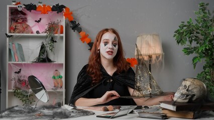 Young woman beauty blogger in scary cape at home studio films video tutorial for halloween party makeup, talks at camera, wears posh witch hat and smiling.