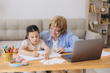 Fototapeta premium Happy grandma helping adorable preschool granddaughter school kid with homework task. Positive school pupil girl and granny reading learning book at laptop together, enjoying education at home