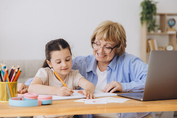 Happy grandma helping adorable preschool granddaughter school kid with homework task. Positive...