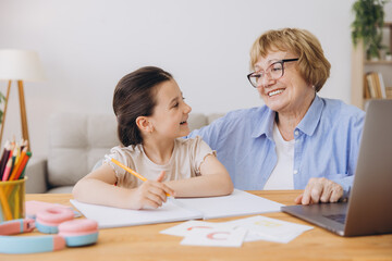 Happy grandma helping adorable preschool granddaughter school kid with homework task. Positive...