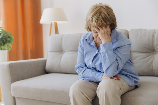 Sick older senior woman sit on sofa feel pain dizziness coping with morning headache concept, upset tired old woman touching sore head suffer from terrible migraine mental problem