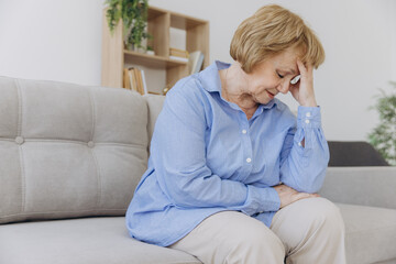 Sick older senior woman sit on sofa feel pain dizziness coping with morning headache concept, upset...