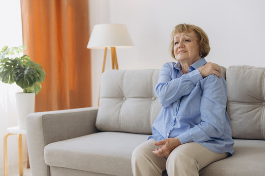 Senior woman sitting on sofa with shoulder pain. Elderly woman is enduring awful ache. Old woman suffering from shoulder injury, health problem concept