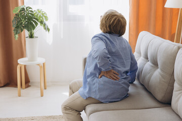 Unhappy old woman touching back, feeling pain, sitting on couch in living room, unhealthy middle...