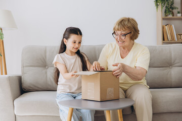 Happy excited grandmother and granddaughter girl opening cardboard box together sits on sofa at...