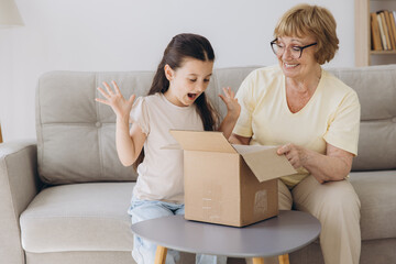 Happy excited grandmother and granddaughter girl opening cardboard box together sits on sofa at home. Shocked family consumers unpack good parcel looking inside great purchase delivered.