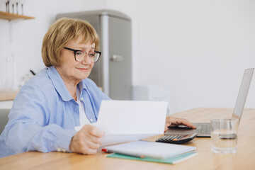 Senior business woman holding paper bill using calculator, old lady managing account finance, calculating money budget tax, planning banking loan debt pension payment sit at home kitchen table.