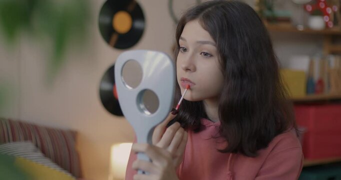 Pretty teenage girl applying lipstick putting on make-up and looking at mirror smiling enjoying beauty sitting in bedroom at home. Fashion and style concept.